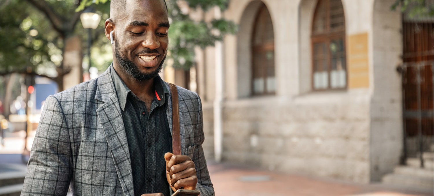 [Featured image] A master's degree student in a grey and black striped jacket and button-up shirt looks at his cellphone while walking across a college campus.