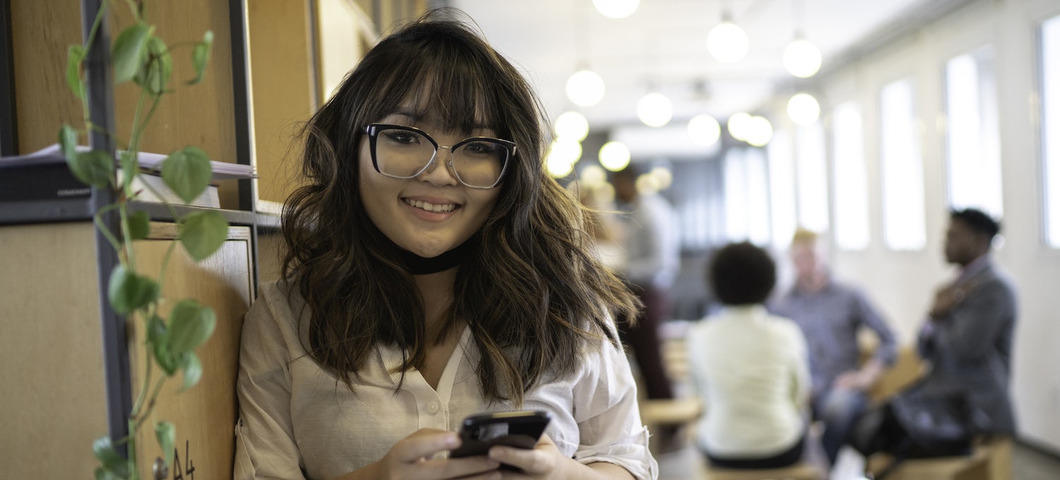 [Featured image] A young person with long, brown hair and glasses stands holding their cell phone and smiling into the camera. 