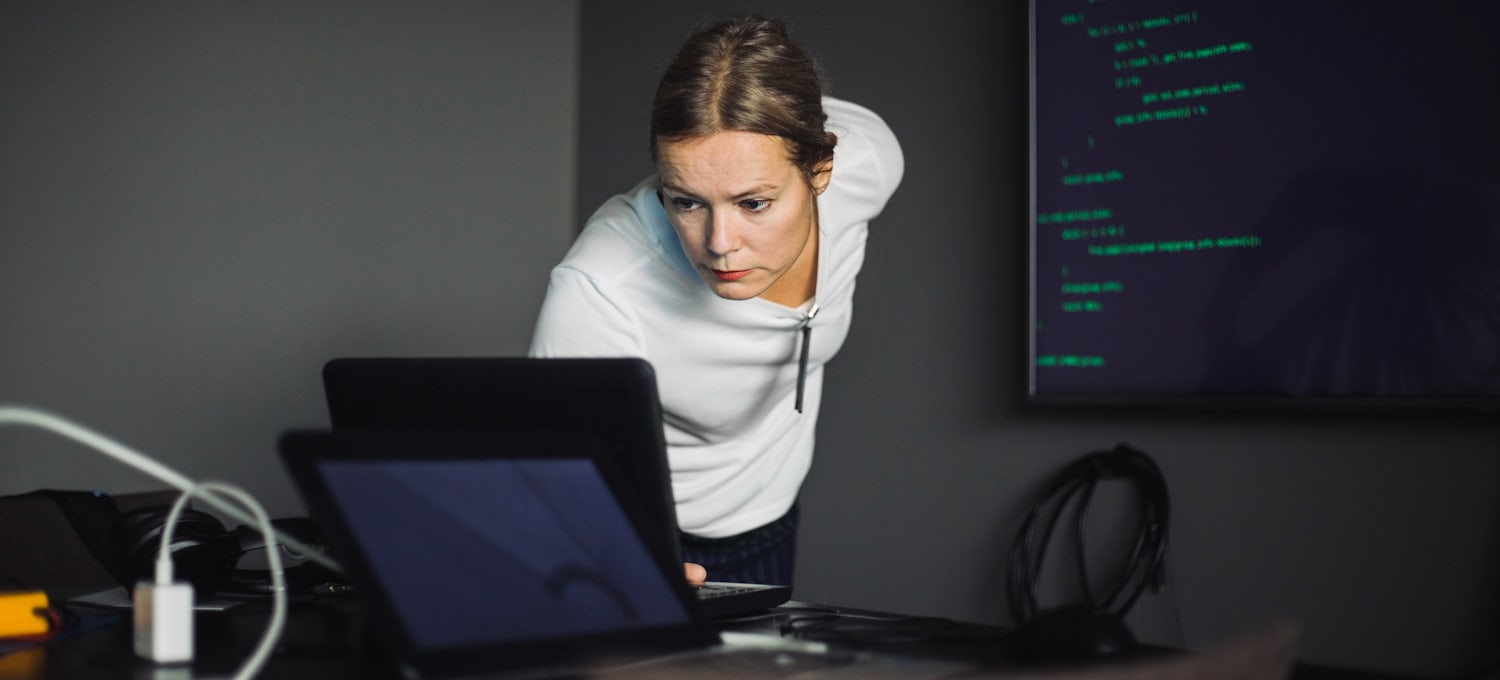 [Featured image] A cybersecurity analyst in a white shirt leans over their desk to view their laptop screen. They are in a dark office and a large monitor with green code is on the wall behind them.