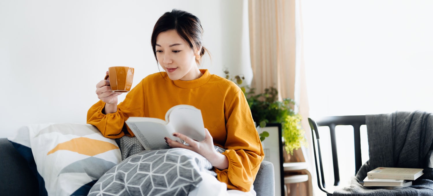 [Featured Image] A person in a mustard-coloured sweater is sitting on their couch drinking coffee while reading a book.