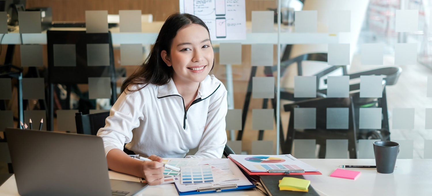 [Featured image] A young professional studies printed charts and data at a desk in front of her laptop and various notebooks to continue learning her craft through upskilling.