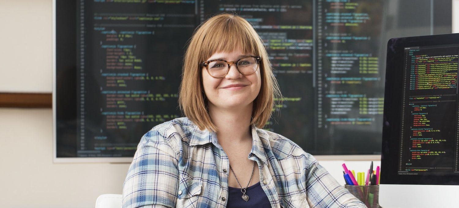 [Featured image] Woman sitting in front of a computer and large monitor smiles after pursuing an alternative to college and finding a coding job.