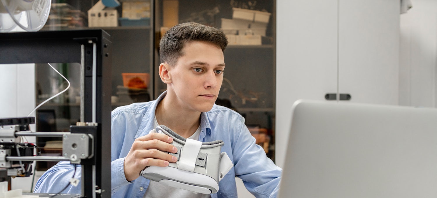 [Featured Image]:  A male, wearing a blue shirt, and a white shirt.  He is sitting in front of his computer screen and holding white goggles. 
