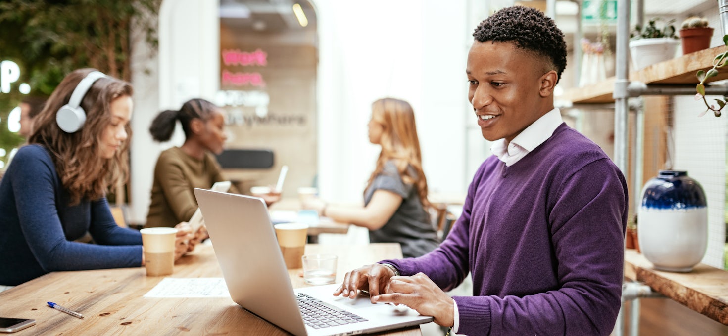 A young man wearing a purple sweater works on his laptop at a shared working station. Three people sit in the background.