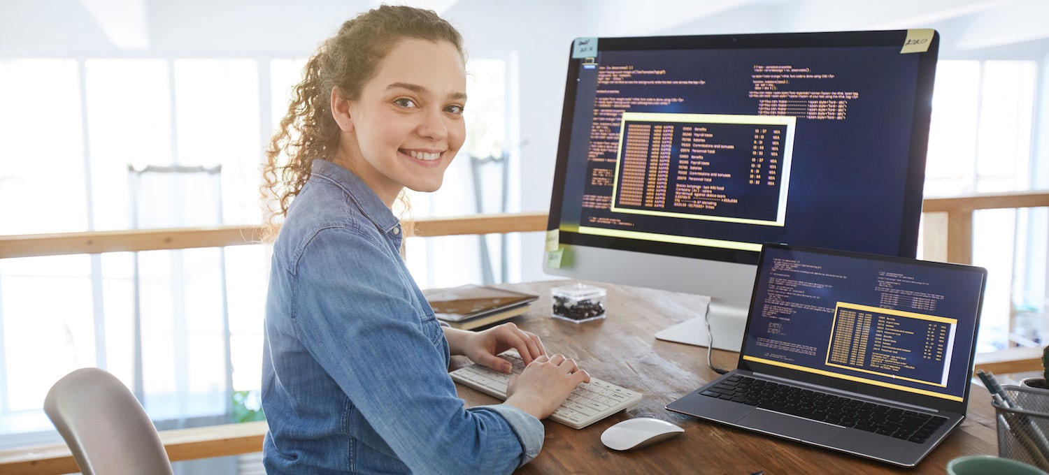 [Featured image] A young person with long curly hair in a ponytail sits in front of a desktop and laptop. They're turned, smiling at the camera. 