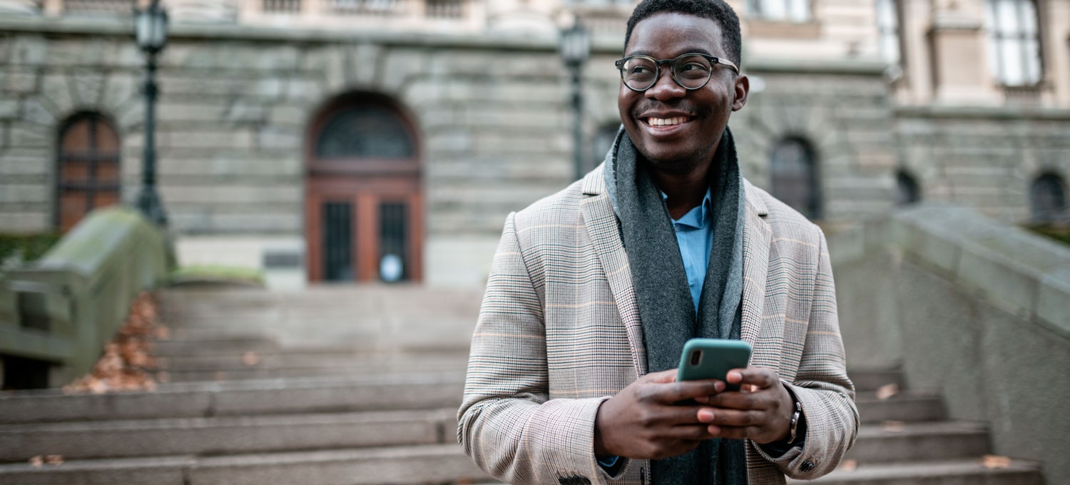 [Featured image] A Doctorate of Business Administration student stands on the steps of a university building while using his cell phone. He's wearing a jacket and scarf.
