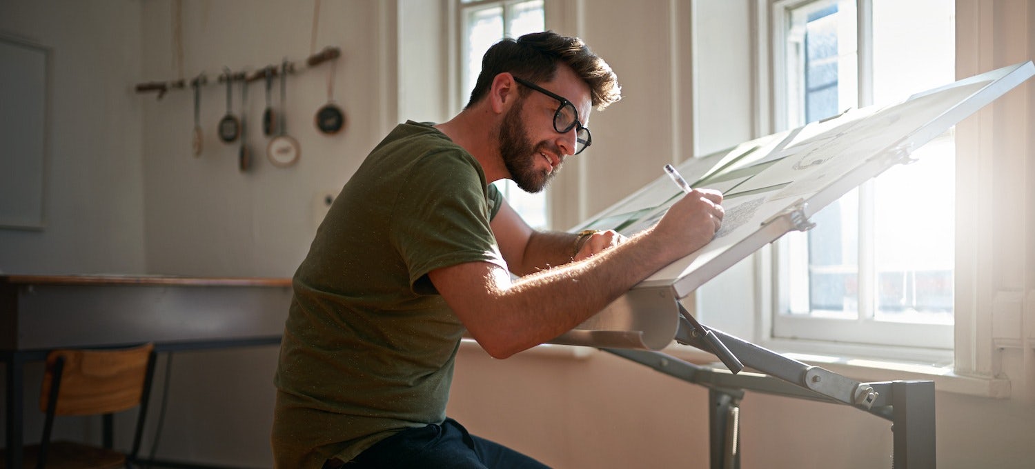 [Featured image] A graphic designer in a green shirt and glasses draws on paper arranged on a drafting table positioned in front of a sunny window.