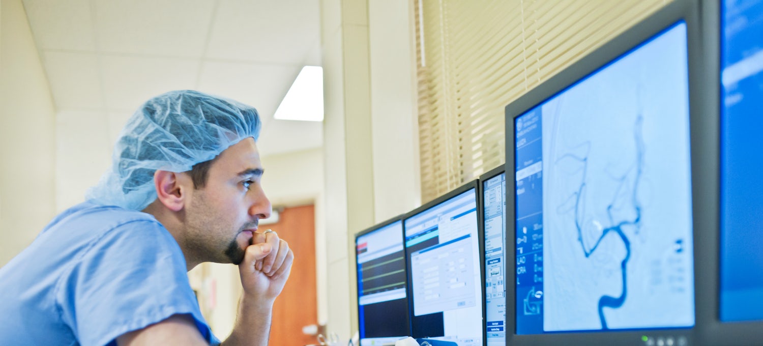 [Featured Image]:  A case manager, wearing a blue uniform and a blue head covering, is sitting in front of three computer screens, looking at charts. 