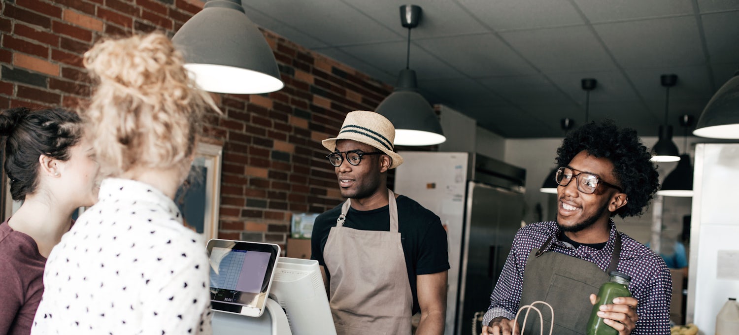 [Featured Image]  Two men, both wearing glasses and aprons, serve their customers at their business.