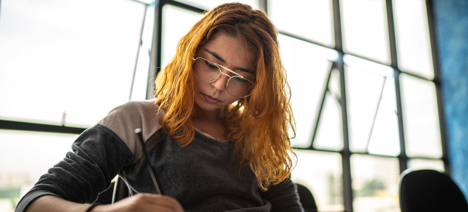 [Featured image] A young woman with orange hair and glasses concentrates as she writes. 