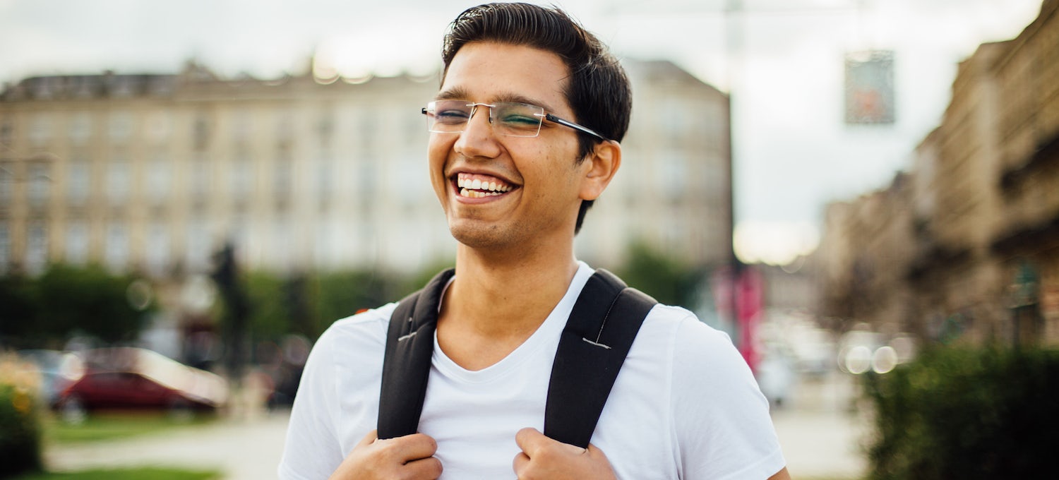 [Featured image] A young Southeast Asian man in glasses and a light blue shirt stands smiling and gripping the straps of his backpack. 