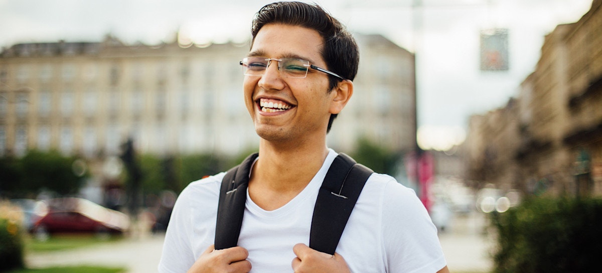 [Featured image] A young Southeast Asian person in glasses and a light blue shirt stands smiling and gripping the straps of their backpack. 