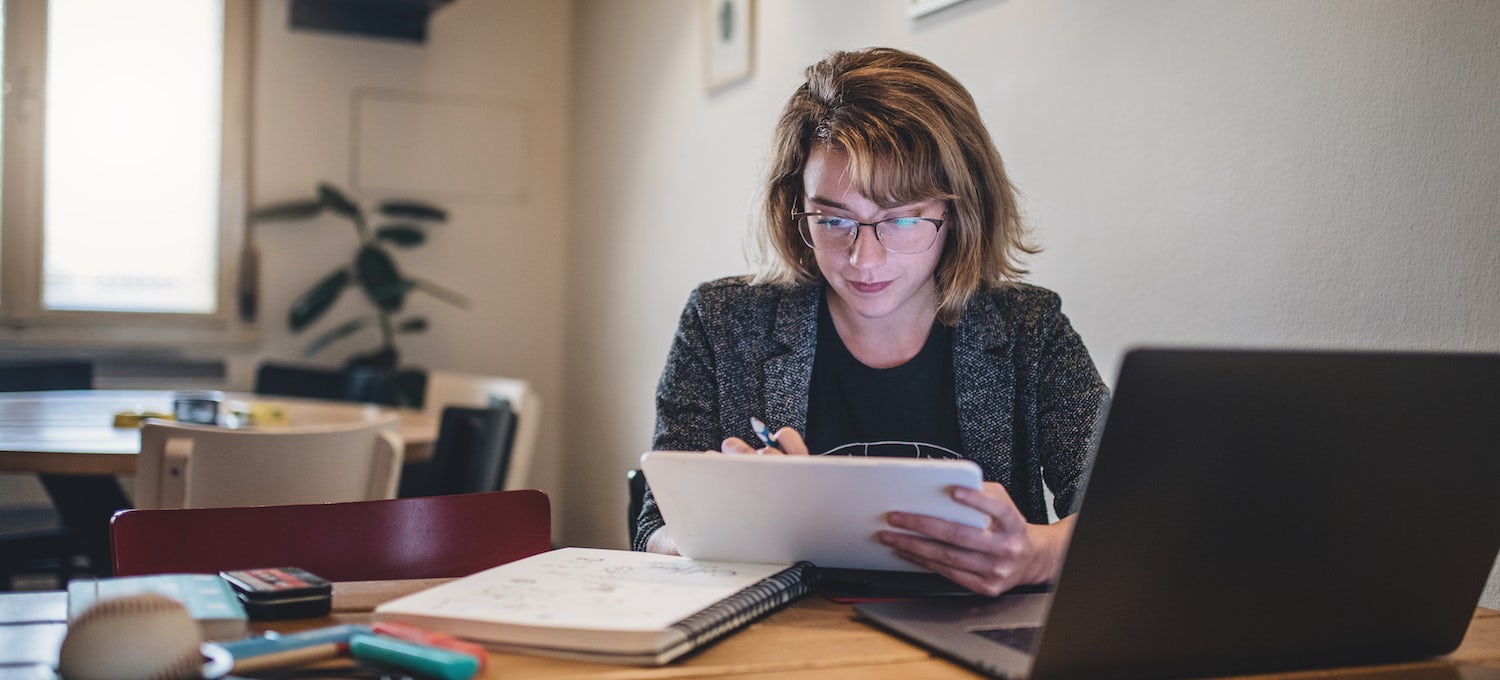[Featured image] A young white woman with glasses stares at a tablet, while her laptop and other paper notebooks are scattered around her.