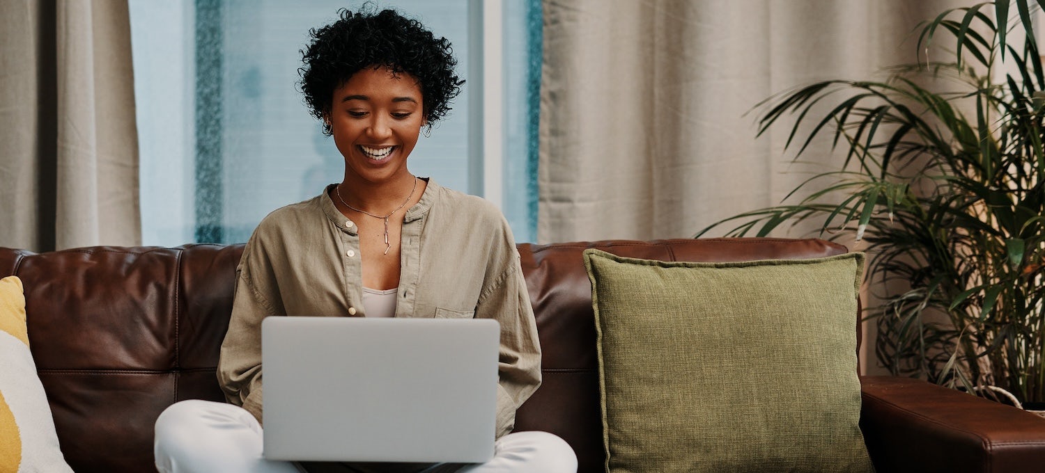 [Featured image] A young woman with short, curly black hair sits on her couch, smiling at her laptop.