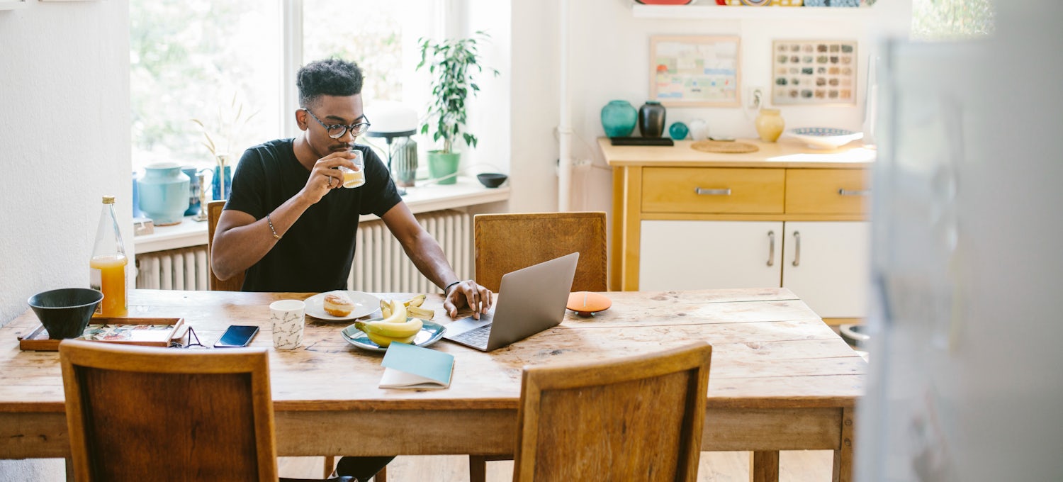 [Featured image] A man sits at his dining room table working on his laptop and drinking a cup of coffee.