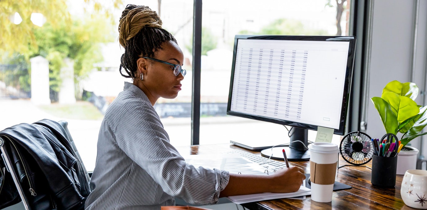 [Imagen destacada] Una mujer con gafas y camisa de rayas se sienta frente a una computadora que busca trabajos de licenciatura en empresariales mientras escribe en sus notas.
