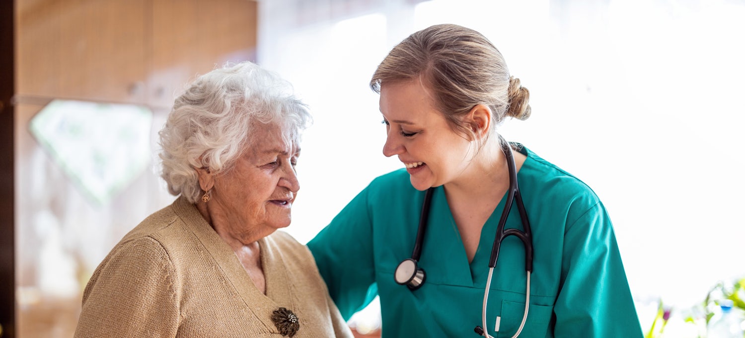 [Featured Image]: A psychiatric nurse wearing a green uniform, and a stethoscope around her neck is taking care of a patient with short white hair and wearing a brown blouse. 