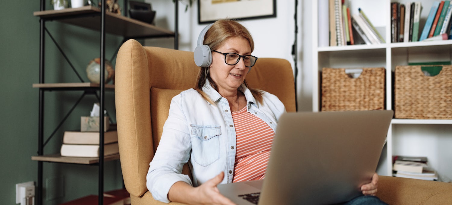 [Featured Image] A person sits in an armchair and chats using a laptop and headphones.