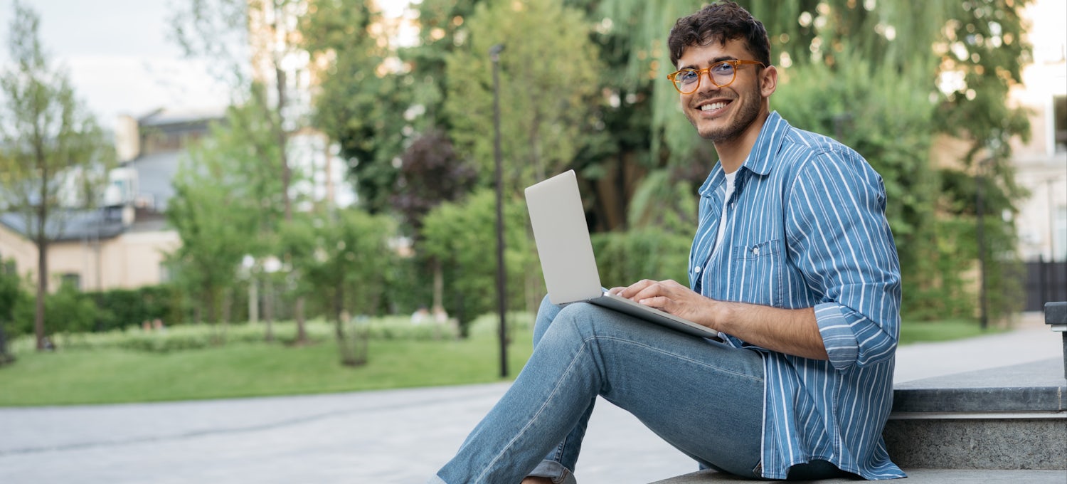 [Featured Image] An interdisciplinary studies degree student in jeans and a blue striped shirt sits outside on steps with a laptop.