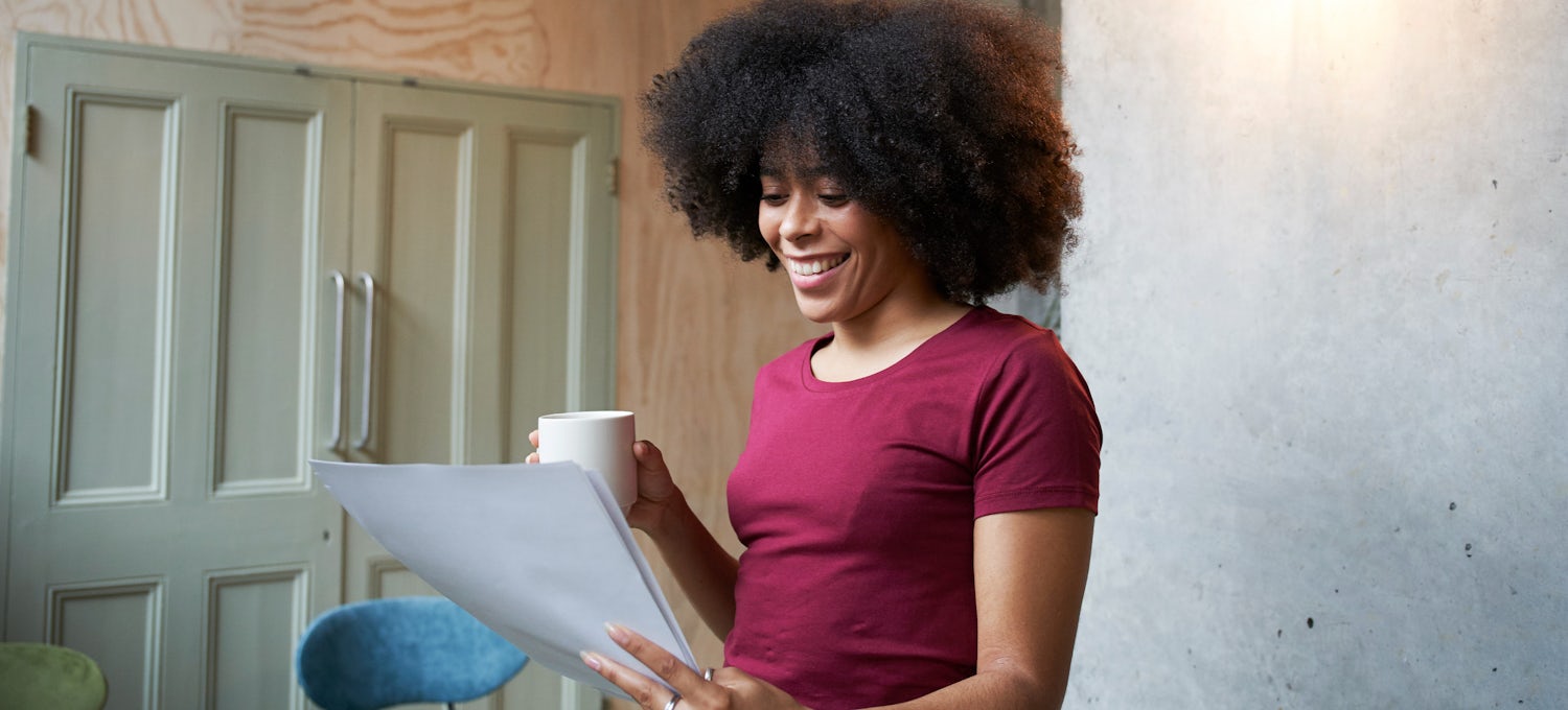 [Featured image] A woman in a maroon shirt holding a cup of coffee looks over her cover letter and smiles.