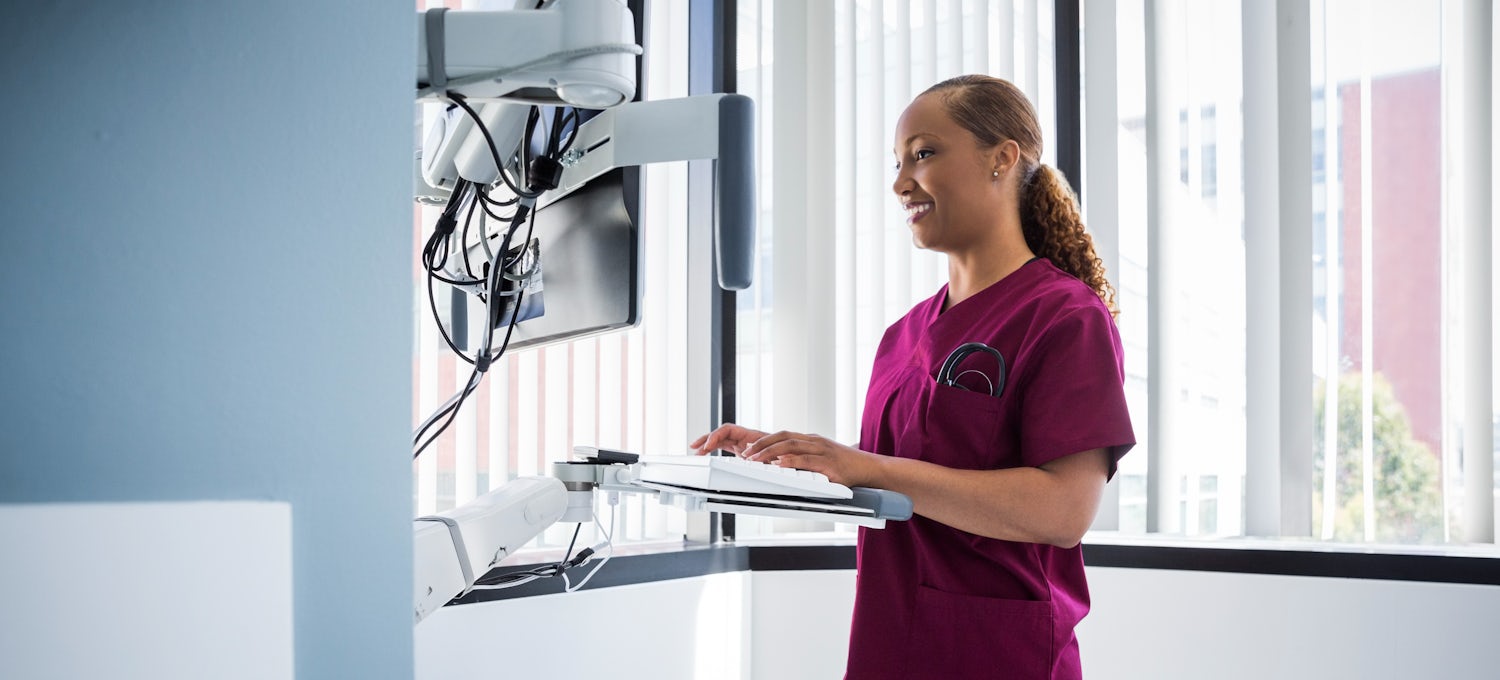 [Featured image] A nursing informatics specialist in maroon scrubs accesses a patient database at a standing workstation in a hospital.