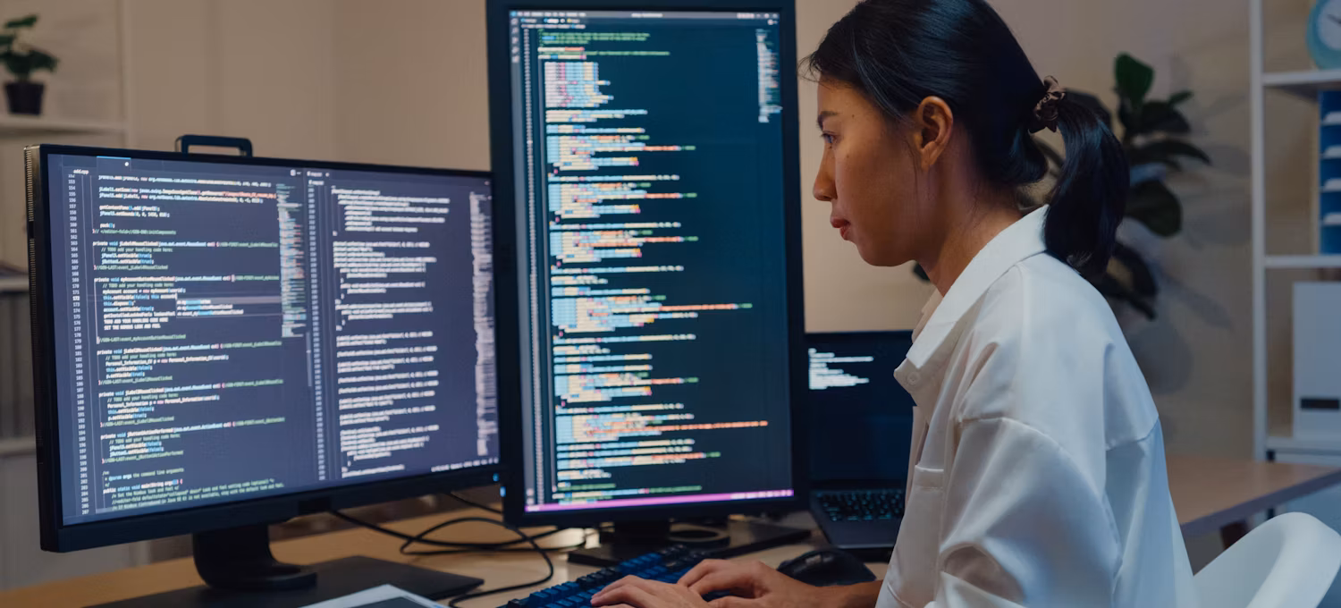 A software developer using a computer to write code sitting at a desk with multiple screens.