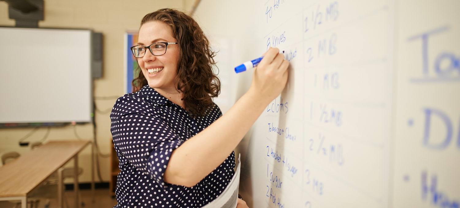[Featured Image] A teacher writes on the whiteboard in her classroom after earning her degree.