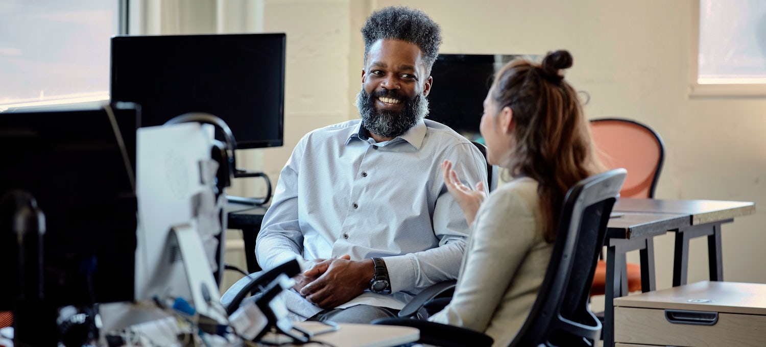 [Featured Image] A bearded man in a grey shirt speaks to a younger woman with a hair bun and a grey suit jacket. 