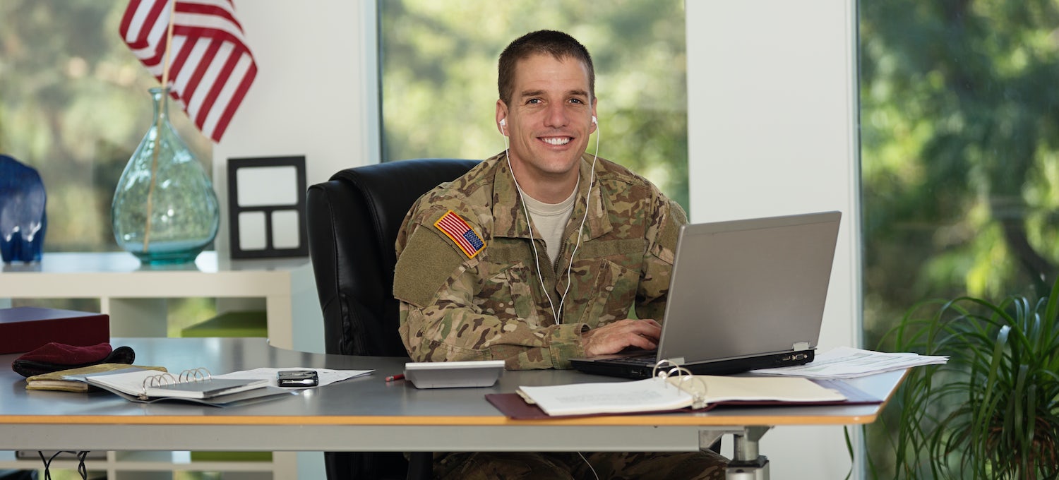 [Featured image] A cryptanalyst wearing army fatigues and white earphones works on a laptop at a desk in front of three windows. 