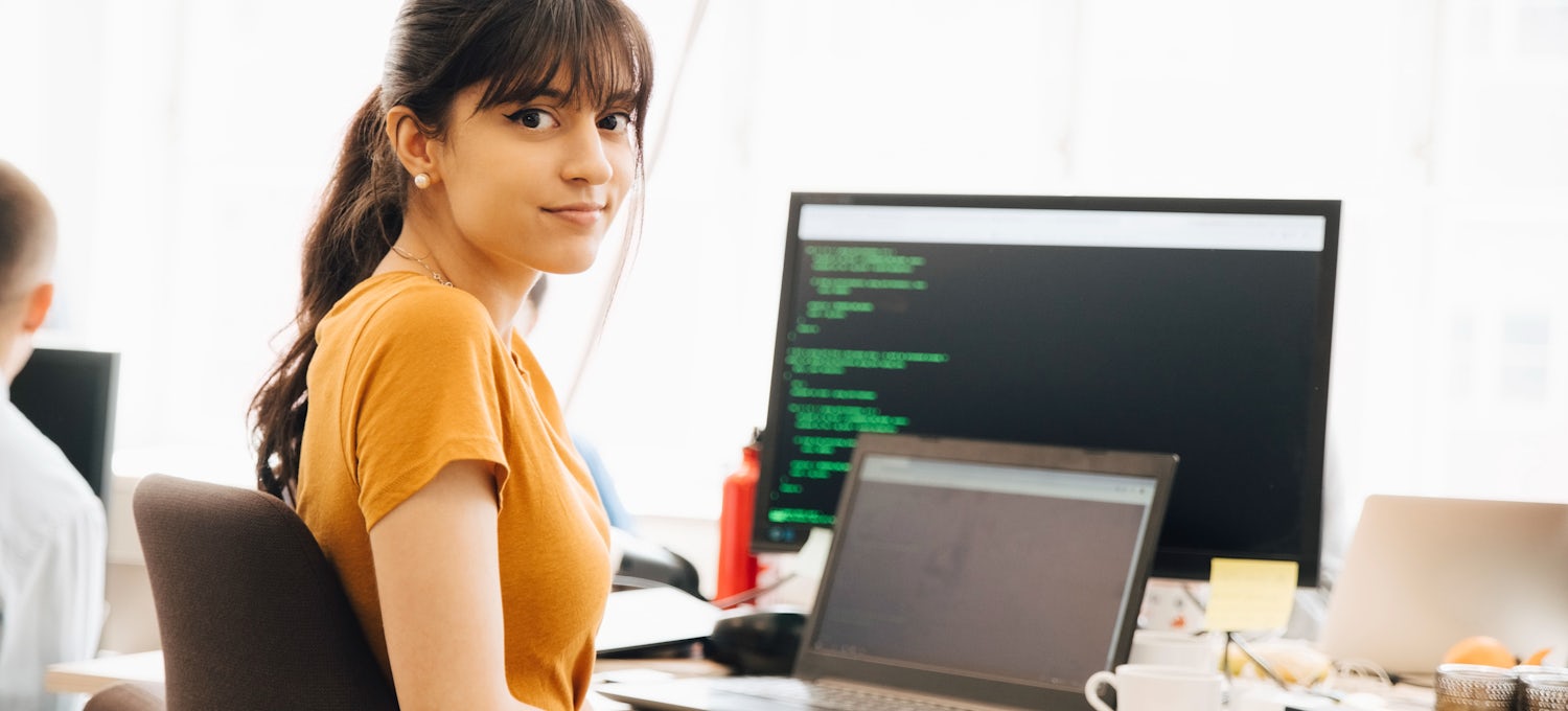 [Featured image] A cybersecurity analyst works with a laptop computer hooked up to an external monitor with green code on a black background running on the screen.