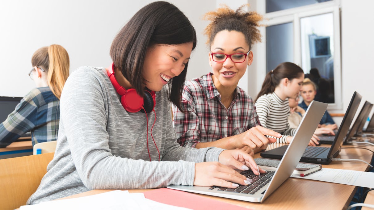 Two women practice writing Python code on their laptops in a classroom lab setting.