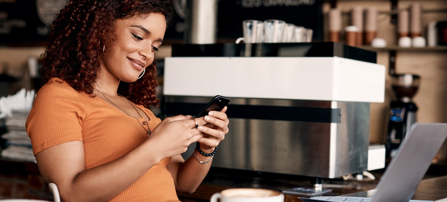[Featured image] A woman in a coffee shop receives a push notification on her cell phone from a geofencing marketing strategy.