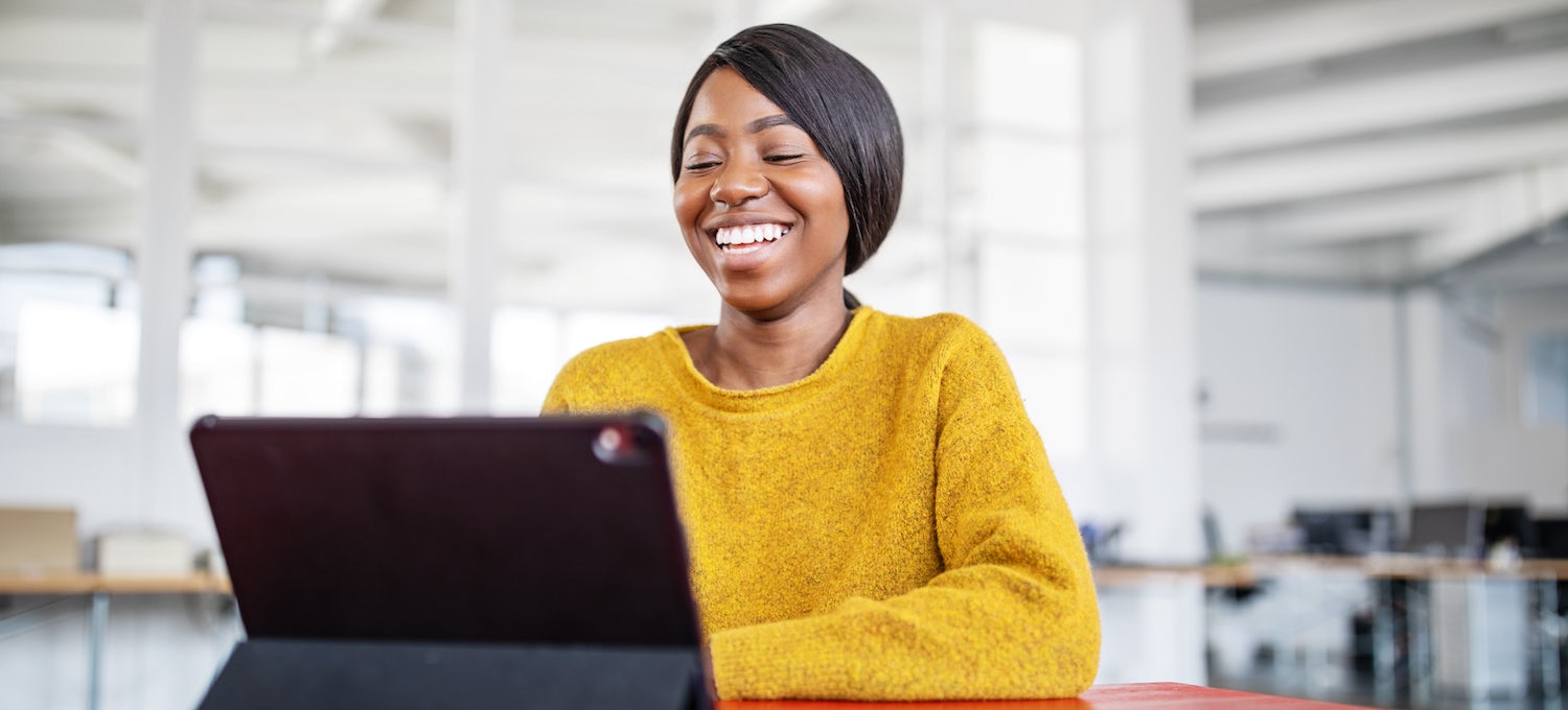 [Featured image] Job candidate smiles at her tablet as she logs on for a video interview with a hiring manager in an open office space.