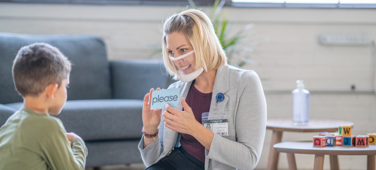 [Featured Image] A blonde, female speech pathologist works with a young male student using flashcards. 