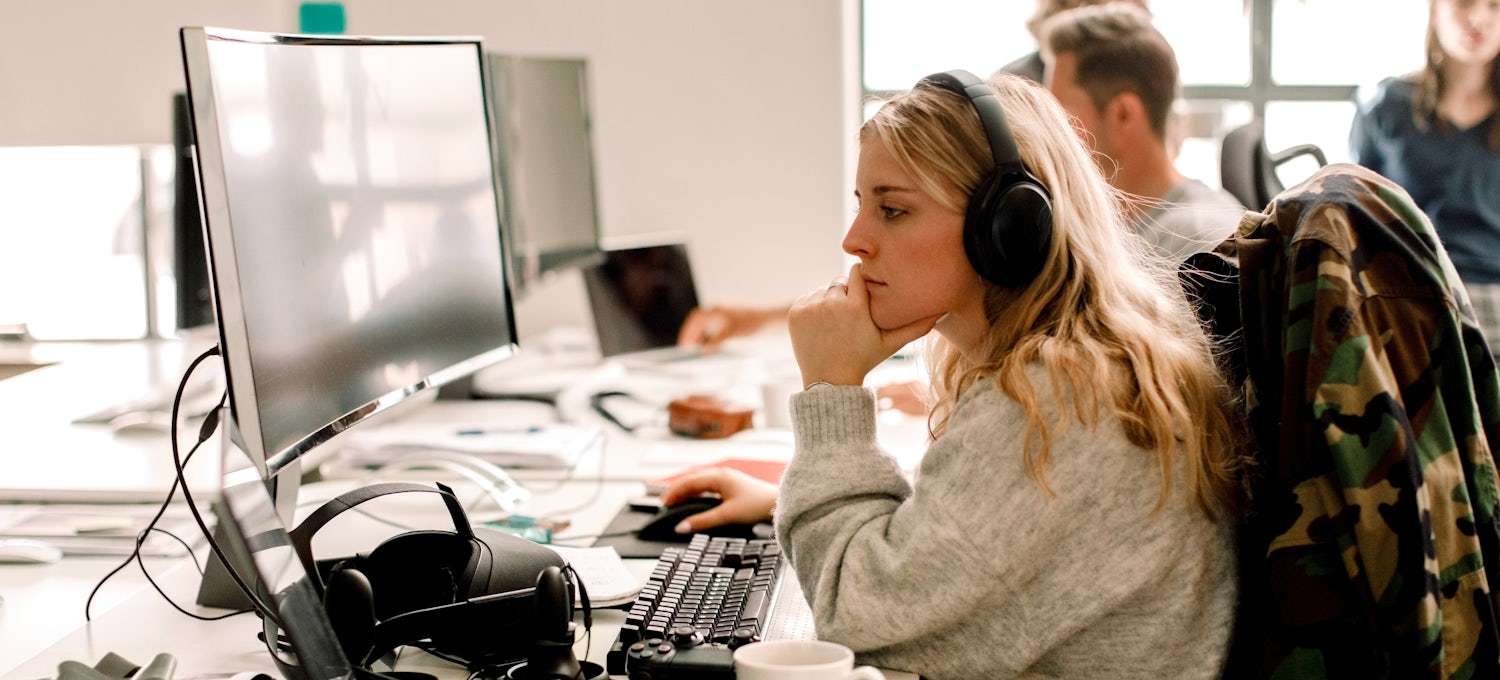 [Featured Image] A woman wearing headphones works on a desktop computer. 