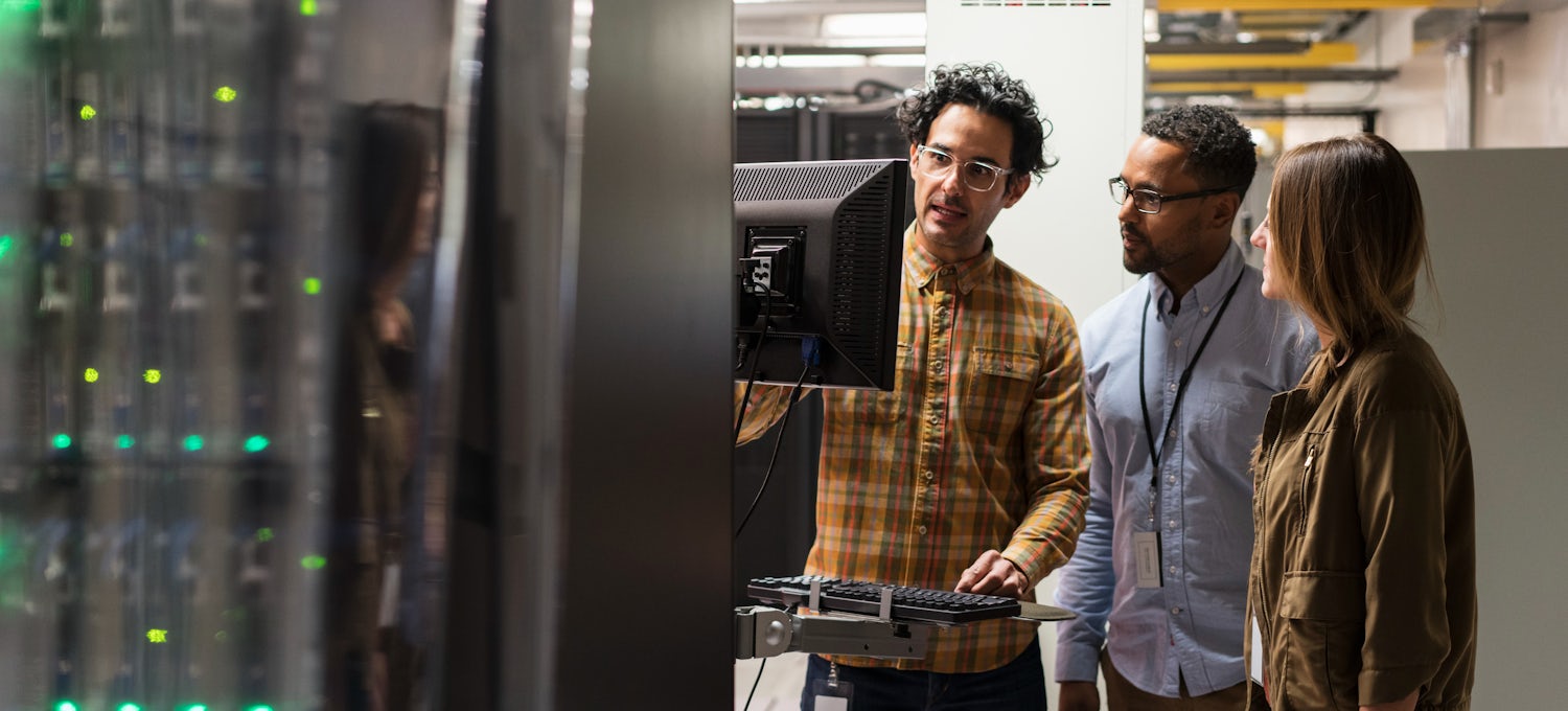 [Featured image] Three cybersecurity analyst colleagues review data on a monitor in a server room.
