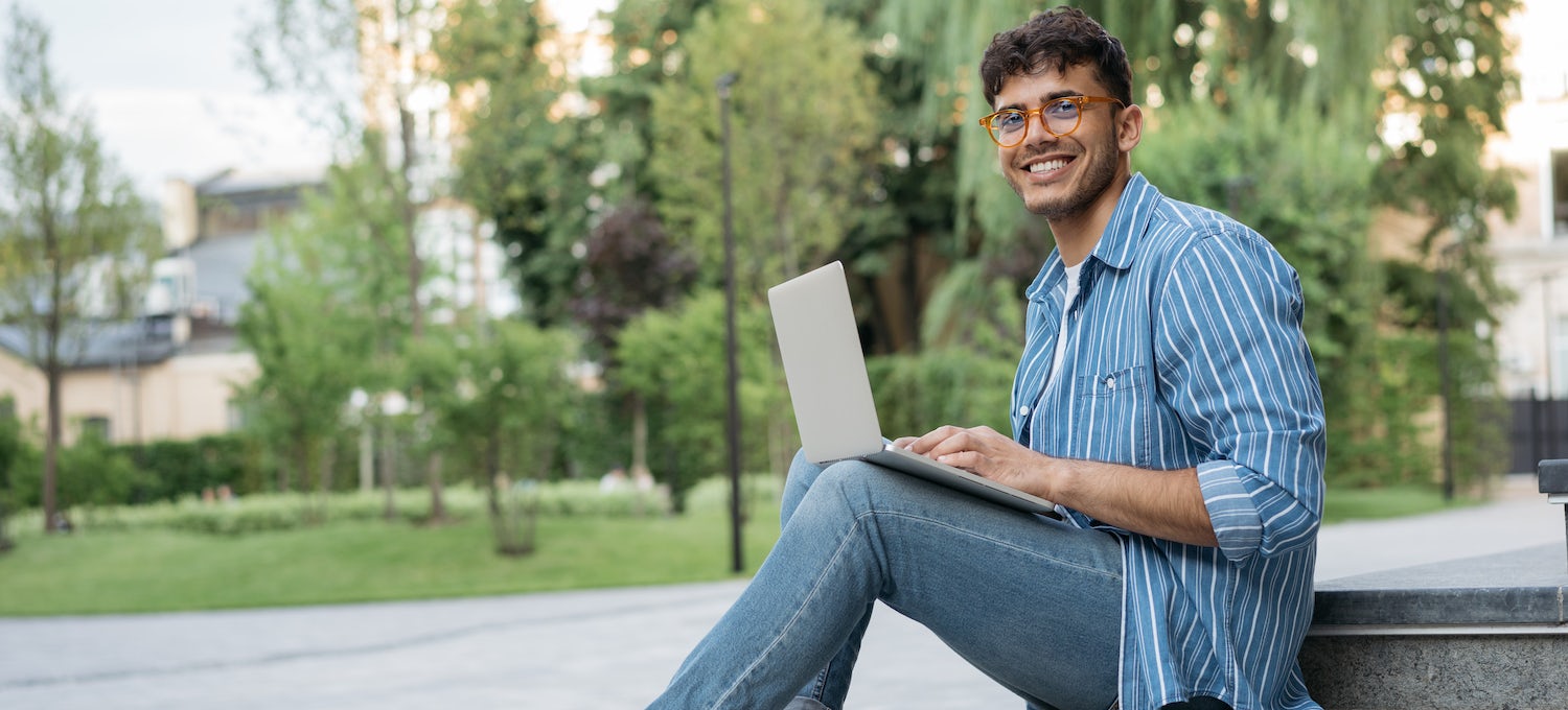 [Featured image] A young man wearing glasses sits with his laptop outside on some steps. 