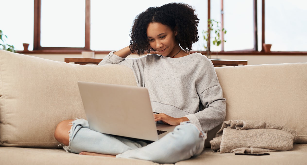 A young Black woman sits on a couch using her laptop. 
