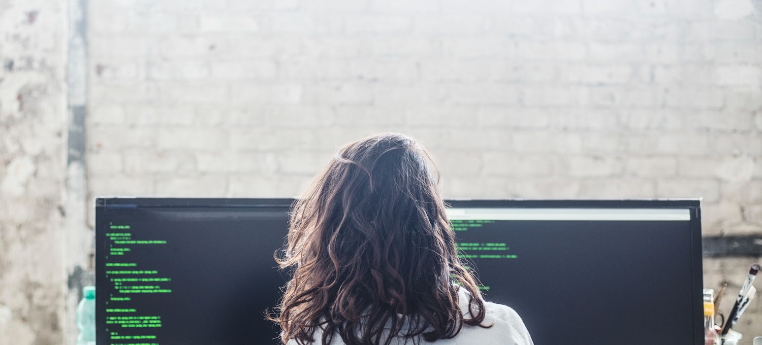 [Featured image]: A female, with long hair and wearing a white jacket, works on two desktop screens in her office, facing a white brick wall,  as she performs her duties as a cloud developer. 