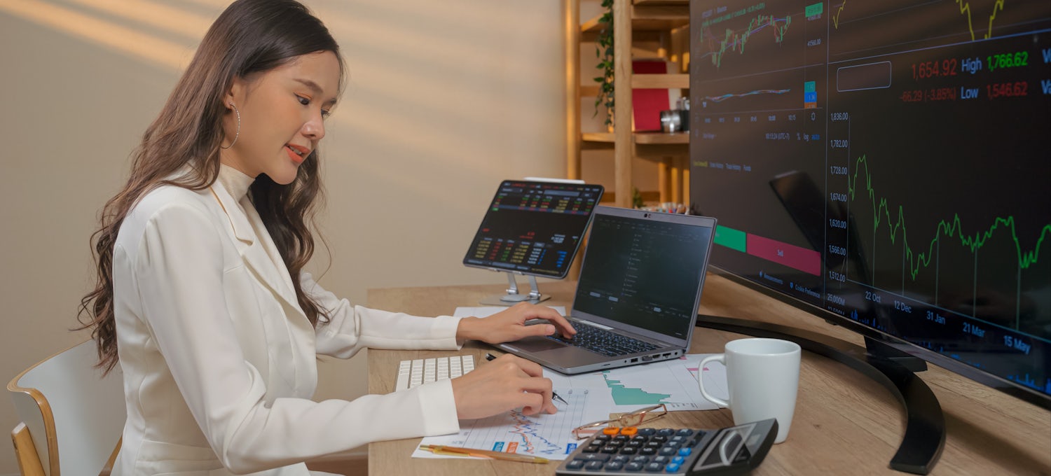 [Featured Image] A businesswoman is sitting at her desk with multiple monitors displaying graphs and trends created using DAX to analyze her data. 
