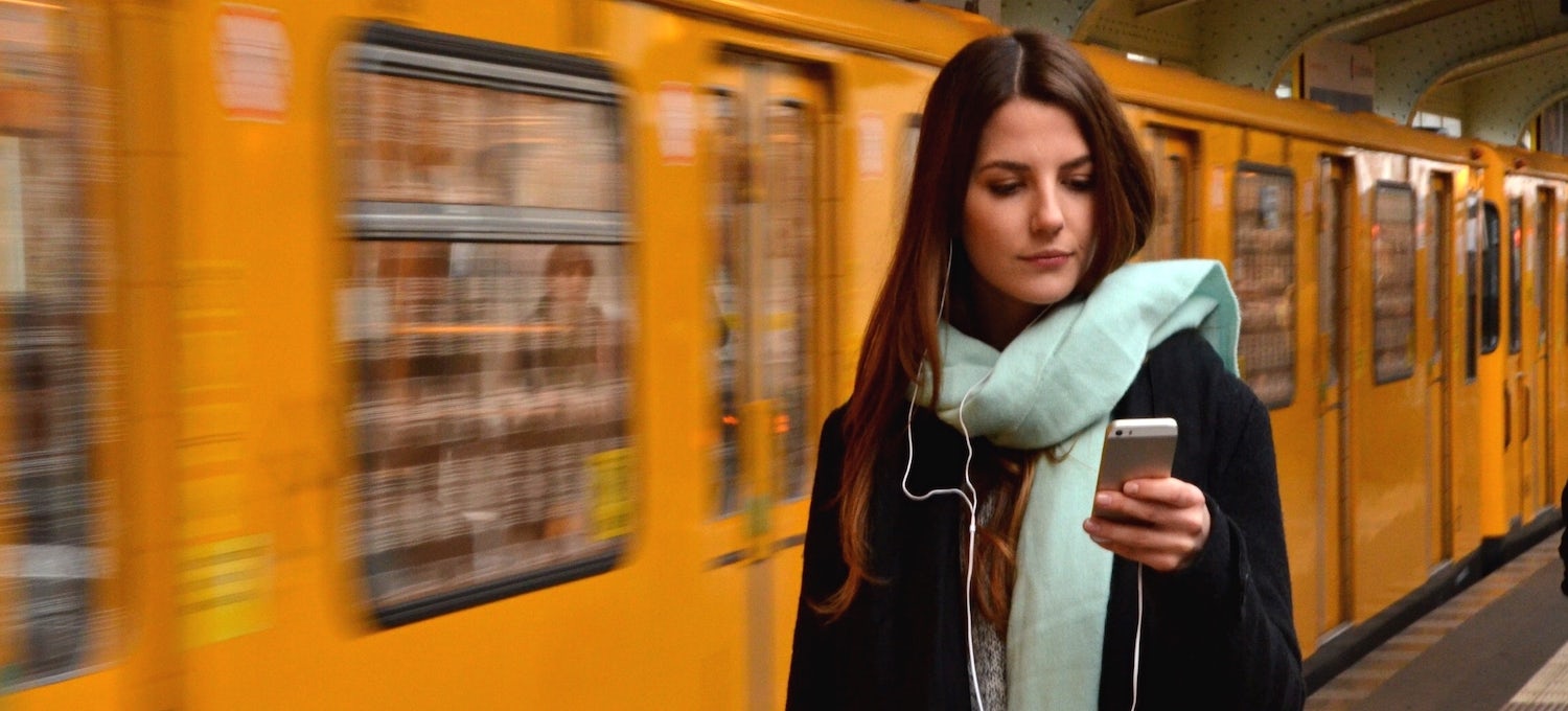 [Featured image] A young woman wearing a black jacket and a light blue scarf stands in front of a yellow train looking at her phone. 