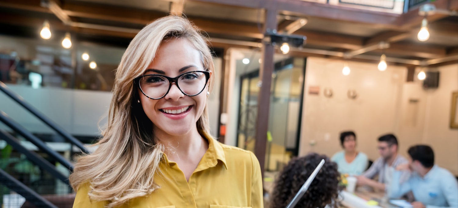 [Featured image] A female with blond hair,  wearing a yellow top, and glasses, is holding her laptop as she prepares for a meeting to discuss promoting products for the international market with the team. 