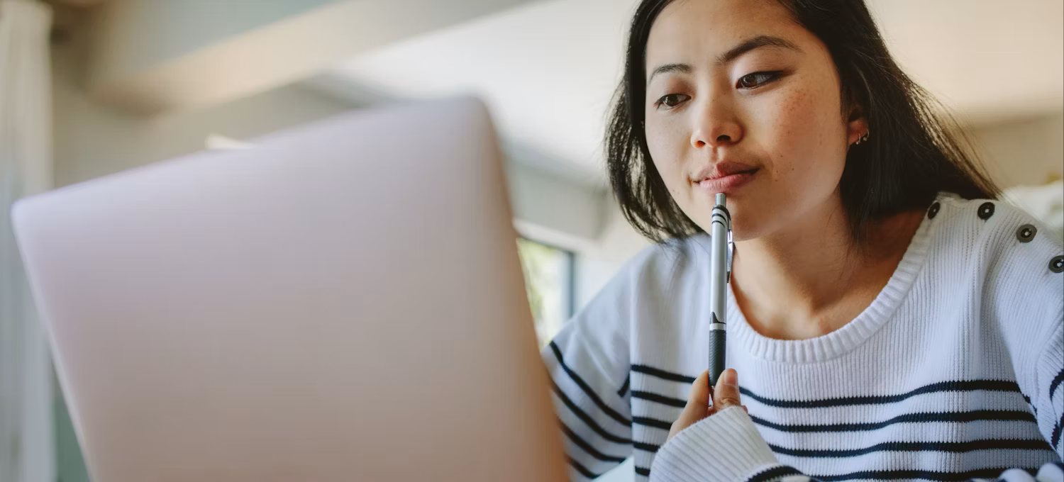 A young woman holding a pen to her lips ponders information she's reading on a laptop.