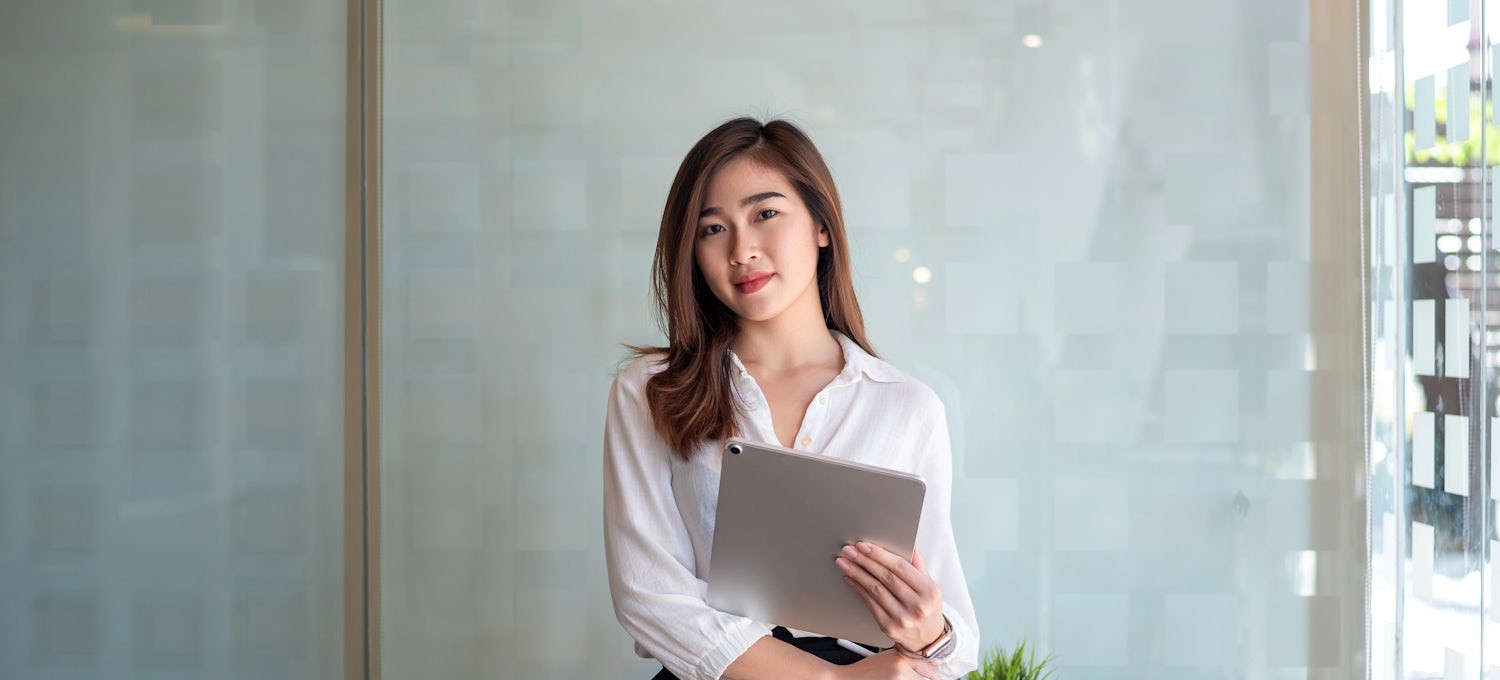 [Featured Image] A project coordinator in a white blouse holds a tablet and stands in her office in front of a frosted glass wall. 