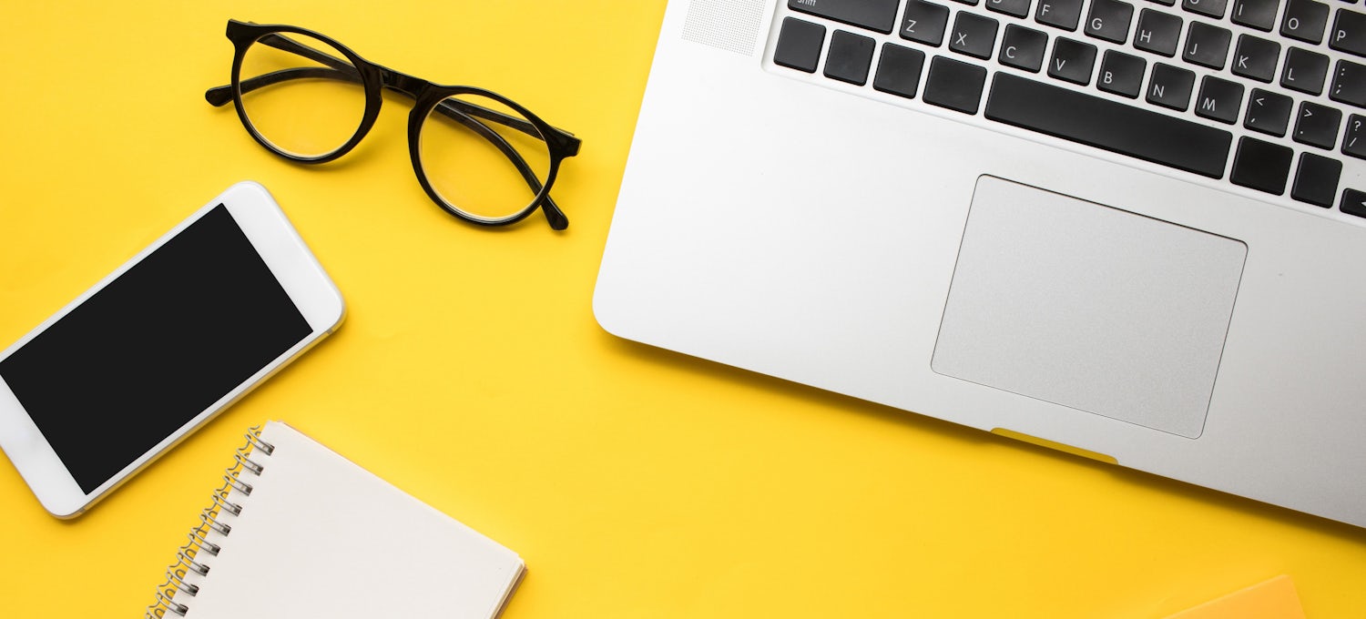 [Featured Image] A laptop keyboard sits on a yellow background with a smartphone and folded pair of glasses beside it.