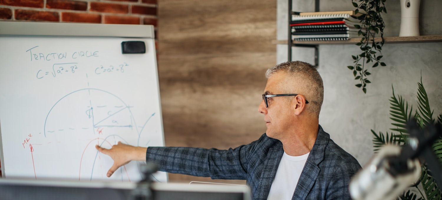 [Featured Image]:   A man wearing a blue plaid shirt and glasses, is standing in front a whiteboard, going over a machine learning project.