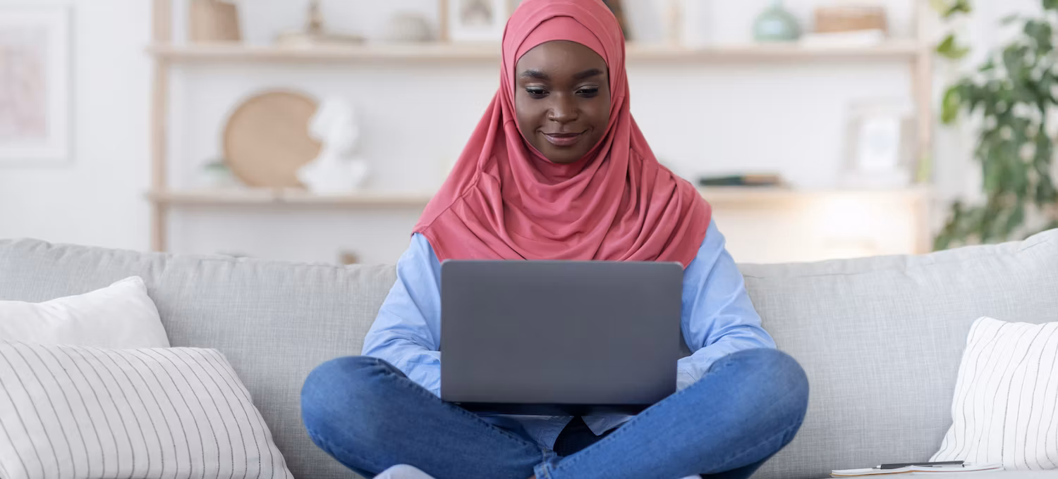 Woman wearing a pink al-amira sits on a grey sofa working on a laptop computer