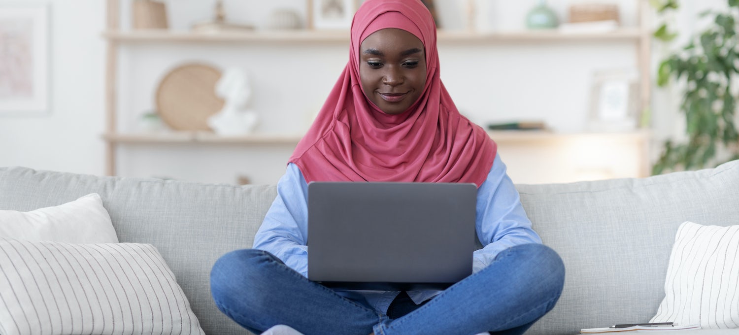 Woman wearing a pink al-amira sits on a grey sofa working on a laptop computer