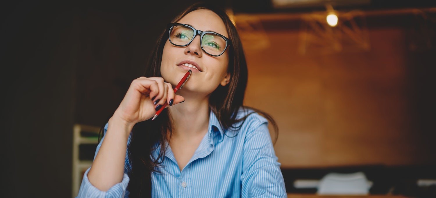 [Imagen destacada] Una mujer con espejuelos, camisa azul, y cabello marrón, sentada frente en un escritorio, reflexionando en la distancia mientras lleva su bolígrafo a su boca.