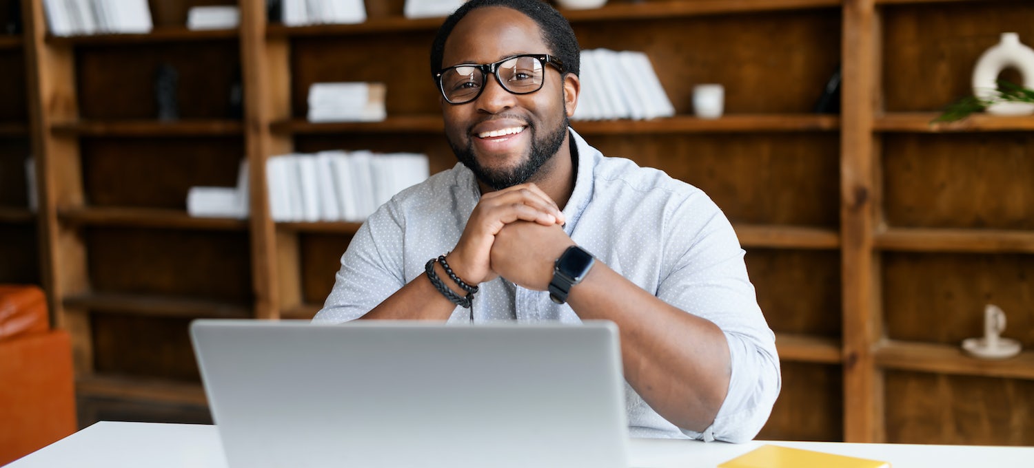 A web designer sits at his laptop computer. He's folding his hands in front of him and smiling at the camera.